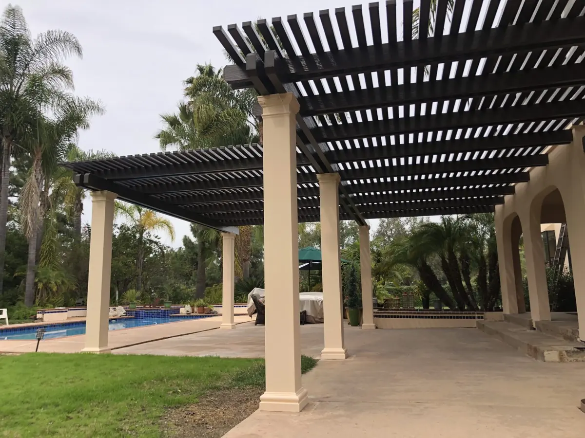 Dark lattice cover on tan columns next to a pool with palm trees